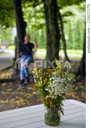 A beautiful bouquet of wildflowers in a glass jar against the background of a park and a silhouette of a young woman on a swing 110072179