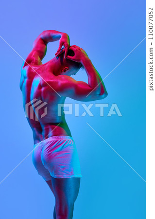 Rear view of shirtless male model with relief strong back, muscular body standing in pants against gradient blue studio background in neon light Rear view of shirtless male model with relief strong back, muscular body standing in pants against gradient blue studio background in neon light 110077052
