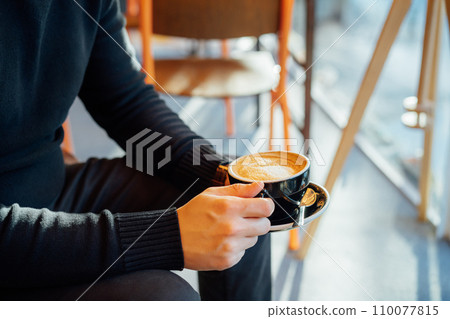 No face handsome young man in black clothes holding hot latte art or cappuccino coffee cup in modern cafe shop. Warm and cozy fall or winter moments. Take a break to relax. Soft selective focus 110077815