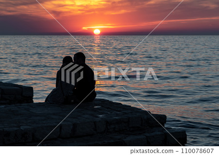Couple Of Young Adults Sit At The Beach Of The Mediterranean Sea And Looks At The Sunset Near The City Of Porec In Croatia 110078607