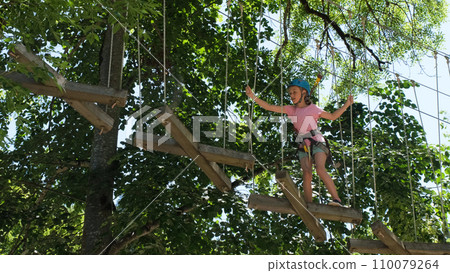 Little girl equipped with safety straps and blue protective helmet climbing on high rope trail in extreme adventure park Little girl equipped with safety straps and blue protective helmet climbing on high rope trail in extreme adventure park 110079264