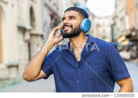 Happy young adult man in wireless headphones choosing, listening music dancing outdoors city street 110080585