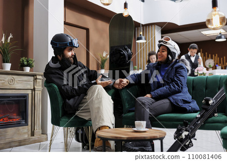 Couple dressed in warm winter gear sitting and talking on cozy sofa in hotel lobby. Excited man and woman wearing ski goggles and winter jackets are holding hands, ski equipment leaning against couch. Couple dressed in warm winter gear sitting and talking on cozy sofa in hotel lobby. Excited man and woman wearing ski goggles and winter jackets are holding hands, ski equipment leaning against couch. 110081406