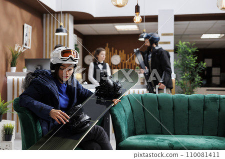 Young female guest carrying and checking her snowboard in lounge area on winter mountain resort. Asian woman seated in hotel reception and maintaining her snowboarding equipment. Young female guest carrying and checking her snowboard in lounge area on winter mountain resort. Asian woman seated in hotel reception and maintaining her snowboarding equipment. 110081411