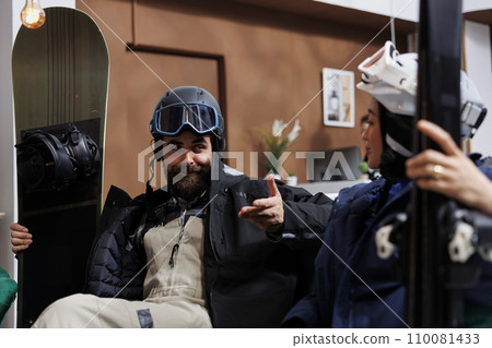 Youthful pair outfitted in cold-weather attire seated on sofa situated in hotel lobby engaged in conversation. Happy man and lady are clutching their wintersports gear set for snowy escapade. Youthful pair outfitted in cold-weather attire seated on sofa situated in hotel lobby engaged in conversation. Happy man and lady are clutching their wintersports gear set for snowy escapade. 110081433
