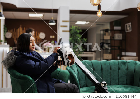 Asian woman relaxes in hotel lobby and prepares her snow gear for wintersports activity. Female tourist with winter jacket grasping her ski goggles skiings skis and helmet in winter mountain resort. Asian woman relaxes in hotel lobby and prepares her snow gear for wintersports activity. Female tourist with winter jacket grasping her ski goggles skiings skis and helmet in winter mountain resort. 110081444