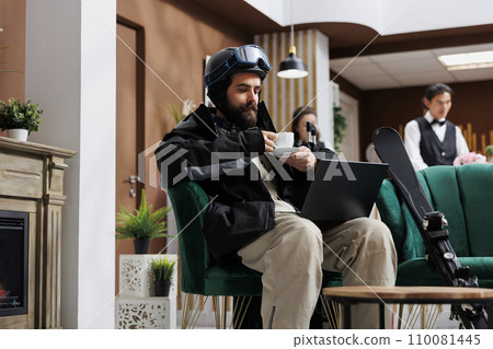 In luxury lounge area, man dressed in winter jacket enjoys coffee while using laptop to explore winter activities. Combination of technology and comfort adds excitement for tourist with skiing gear. In luxury lounge area, man dressed in winter jacket enjoys coffee while using laptop to explore winter activities. Combination of technology and comfort adds excitement for tourist with skiing gear. 110081445