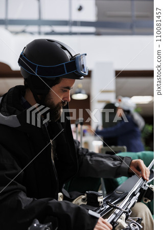 Close-up of caucasian traveler adjusting ski gear in hotel lobby. Male tourist, wearing winter jacket and ski goggles, ensuring safety of wintersports equipment. Ready to explore ski resort for skiing Close-up of caucasian traveler adjusting ski gear in hotel lobby. Male tourist, wearing winter jacket and ski goggles, ensuring safety of wintersports equipment. Ready to explore ski resort for skiing 110081457