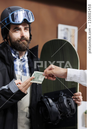 Detailed view of male snowboarder tipping employee with cash in ski hotel reception. Caucasian man in wintersport clothing giving gratuity to concierge at winter mountain resort. Detailed view of male snowboarder tipping employee with cash in ski hotel reception. Caucasian man in wintersport clothing giving gratuity to concierge at winter mountain resort. 110081458