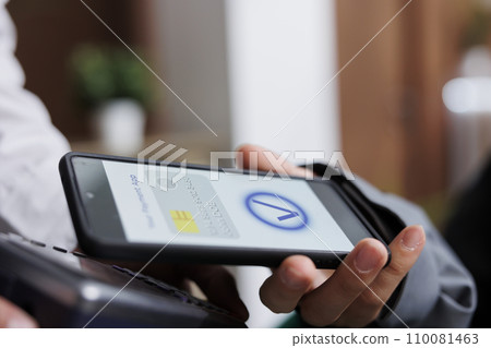 Close-up shot of tourist making contactless payment via mobile phone using NFC technology. Detailed view of pair of hands grasping pos terminal and smartphone, paying winter holiday reservation. Close-up shot of tourist making contactless payment via mobile phone using NFC technology. Detailed view of pair of hands grasping pos terminal and smartphone, paying winter holiday reservation. 110081463