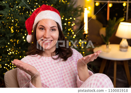A young attractive woman in cozy pajamas is sitting at home on a chair in a red Christmas hat, smiling and spreading her arms to the sides A young attractive woman in cozy pajamas is sitting at home on a chair in a red Christmas hat, smiling and spreading her arms to the sides 110081918
