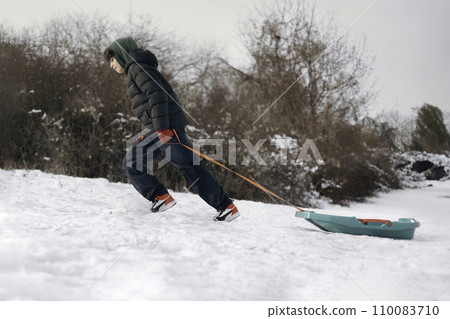 Teenager clad in a winter jacket and hood vigorously pulls a bright teal sled by a red rope across a snow-covered landscape.Concept representing winter sports, childhood, leisure activities during the Teenager clad in a winter jacket and hood vigorously pulls a bright teal sled by a red rope across a snow-covered landscape.Concept representing winter sports, childhood, leisure activities during the 110083710
