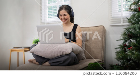 Young Asian woman listening music from headphones and sitting on sofa in living room 110084200
