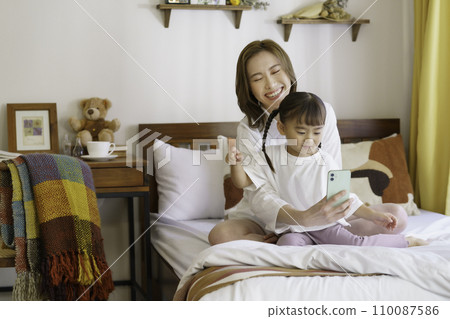 一位年輕的棕髮母親和她 4 歲的女兒在床上操作智慧型手機 110087586