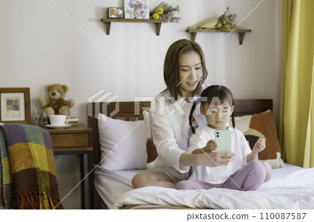 一位年輕的棕髮母親和她 4 歲的女兒在床上操作智慧型手機 110087587