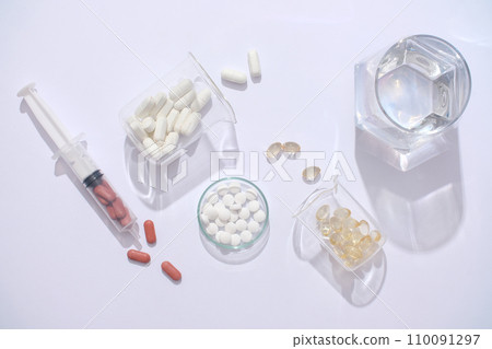 On a white backdrop, a glass of water, syringe, and medicines arranged on a transparent petri dish and lab beaker. Essential aids in supporting the treatment process. 110091297