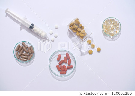 Against a minimalist backdrop, a glass of water, syringe, and medicines are showcased on a transparent petri dish and beaker. Medicine play a crucial role in supporting the treatment process. Against a minimalist backdrop, a glass of water, syringe, and medicines are showcased on a transparent petri dish and beaker. Medicine play a crucial role in supporting the treatment process. 110091299