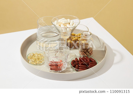 Close-up of a glass of water and a variety of dietary supplements displayed in glassware on a ceramic plate. Dietary supplements are produced in the form of tablets, capsules, liquids, etc. Close-up of a glass of water and a variety of dietary supplements displayed in glassware on a ceramic plate. Dietary supplements are produced in the form of tablets, capsules, liquids, etc. 110091315