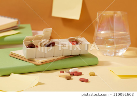 A daily pill box with lots of pills of different sizes and colors. A glass of water and notebook are placed on the table. Copy space with a medical theme. 110091608