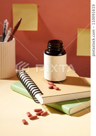 An unlabeled bottle of dietary supplements is placed on top of two notebooks. Yellow sticky notes are stuck on the wall. Mockup for medicine advertising. 110091619