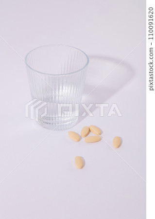 A glass of water and pastel pink pills are displayed on a white background. Minimalist space. Advertising drugs and functional foods. Commercial images. 110091620