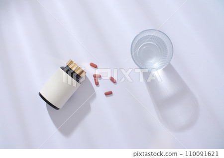 Top view of an unbranded medicine bottle, pills and a glass of water on a white background with drop shadow. Blank label for lettering design. New drug launch. Top view of an unbranded medicine bottle, pills and a glass of water on a white background with drop shadow. Blank label for lettering design. New drug launch. 110091621