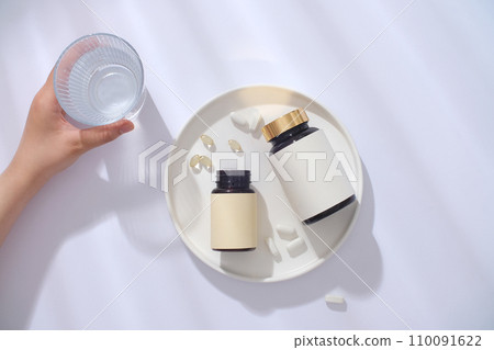Two unlabeled bottles of dietary supplements are displayed on a white ceramic tray. A hand holds a glass of water next to it. Minimalist white background. 110091622