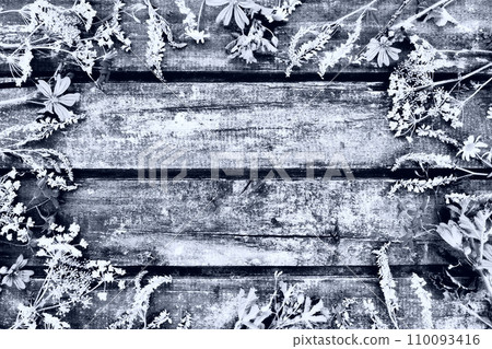 Wildflowers arranged in a circle on a wooden table background. Chamomile, sweet clover, wild geranium, bluebells, parsley inflorescences. Wooden horizontal boards. Monochrome image. 110093416