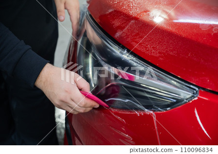 The master applies vinyl film to the headlight of a red car. Closeup view on worker detailer hand smoothing with a scraper protective film.  110096834