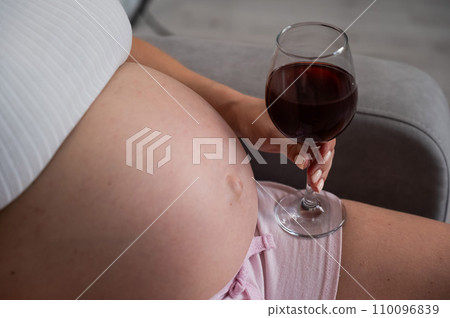 Close-up of the belly of a pregnant woman holding a glass of red wine while sitting on the sofa.  110096839