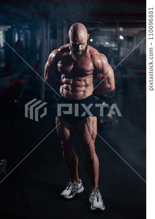 Muscular bald man posing in shorts. Bodybuilder showing off his shape in the gym.  110096881