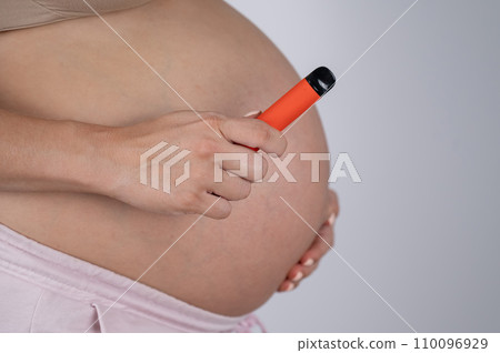A pregnant woman smokes a vape. A girl holds an electronic cigarette against the background of her bare tummy. Copy space. A pregnant woman smokes a vape. A girl holds an electronic cigarette against the background of her bare tummy. Copy space. 110096929