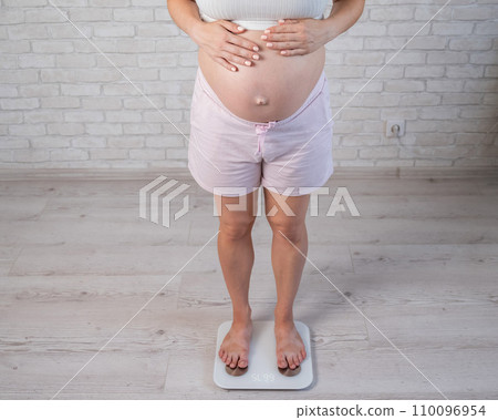 Top view of a pregnant woman in home clothes standing on an electronic scale and holding her hands on her tummy. Top view of a pregnant woman in home clothes standing on an electronic scale and holding her hands on her tummy. 110096954