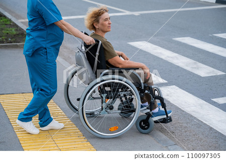 Profile of a nurse helping an elderly woman in a wheelchair cross the road. Profile of a nurse helping an elderly woman in a wheelchair cross the road. 110097305