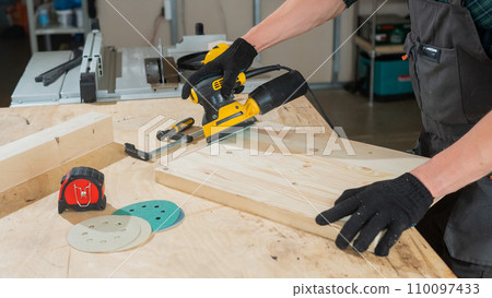 A man using an orbital wood sander in a workshop. Close-up of a carpenter's hands. A man using an orbital wood sander in a workshop. Close-up of a carpenter's hands. 110097433