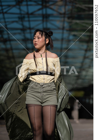 A beautiful Asian woman in shorts and a green leather coat comes out of the subway.  110097652