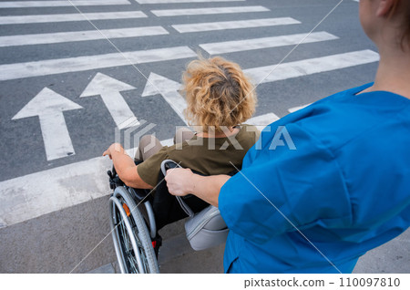 Rear view of a nurse helping an elderly woman in a wheelchair cross the road. Rear view of a nurse helping an elderly woman in a wheelchair cross the road. 110097810