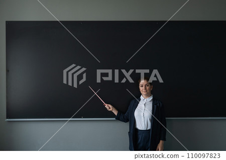 Red-haired caucasian woman in a trouser suit. Smiling female teacher with a pointer at the blackboard.  110097823