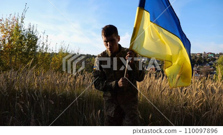 Young male military in uniform walks with waving flag of Ukraine at countryside. Soldier of ukrainian army going with lifting national banner outdoor. Victory against russian aggression concept 110097896
