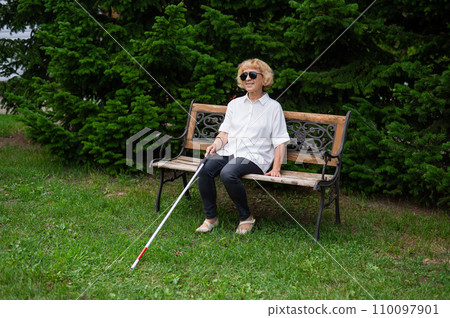 An elderly blind woman sits on a bench in the park. 110097901