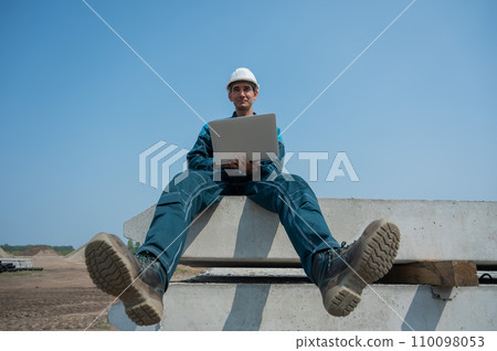 Caucasian male builder in hardhat sits on floor slabs and uses laptop at construction site. Caucasian male builder in hardhat sits on floor slabs and uses laptop at construction site. 110098053