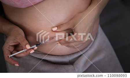 A pregnant woman puts an injection of insulin while sitting on the couch. Close up of the belly. 110098093