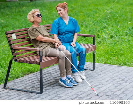 A nurse and an elderly blind woman are sitting on a bench in the park. 110098393