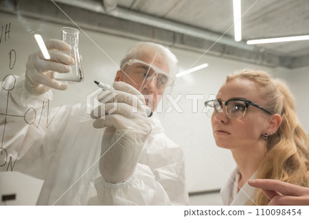 Two chemist colleagues write formulas on glass. Caucasian elderly man and young woman brainstorming. Two chemist colleagues write formulas on glass. Caucasian elderly man and young woman brainstorming. 110098454