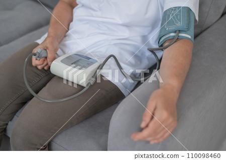 Mature Caucasian woman measuring blood pressure while sitting on sofa at home.  110098460