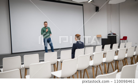 A red-haired Caucasian businesswoman sits in the front row of an empty conference room. Bearded man giving a lecture.  110098461