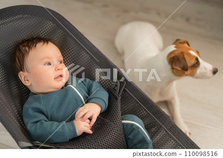 A dog sits next to a cute three-month-old boy dressed in a blue overalls in a baby lounger. 110098516