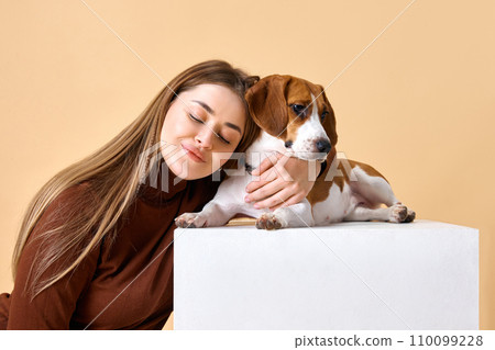 Studio portrait of dog and owner. Young woman smiling and hugging her favorite puppy Beagle dog against beige background. 110099228