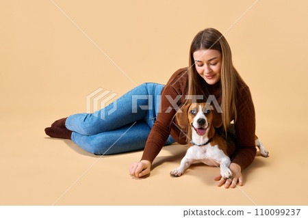 Love between dog and owner. Young woman in casual clothes posing with her favorite pet against beige background. Concept of friendship 110099237