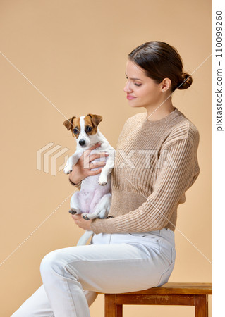 Portrait of young attractive woman sitting on chair and hugs her lovely cute pet against beige studio background. Concept of love. 110099260